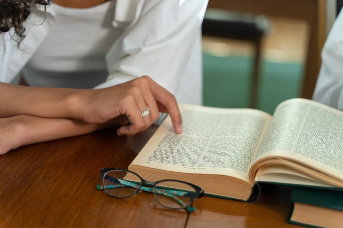 Pessoa lendo livro acadêmico em biblioteca universitária com caderno de anotações ao lado