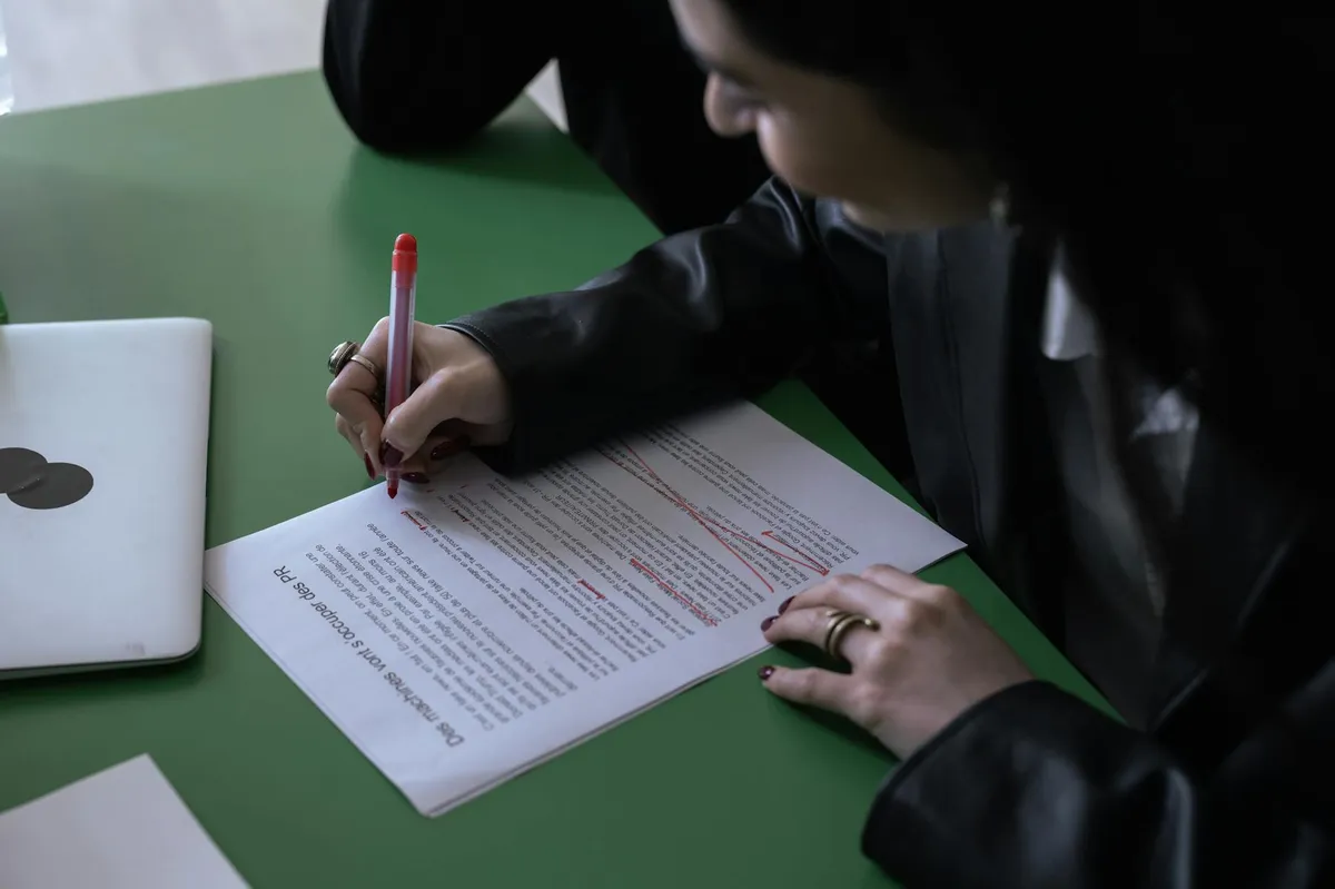 Mão de pesquisador com caneta sobre folhas de texto impresso marcado com anotações em vermelho