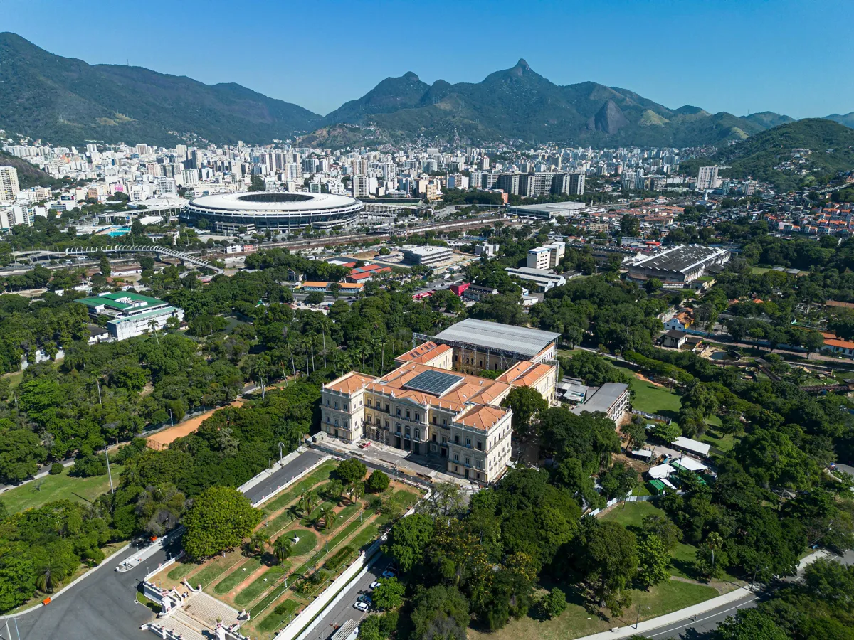 Pesquisadora consultando documentos de edital em mesa com computador e xícara de café