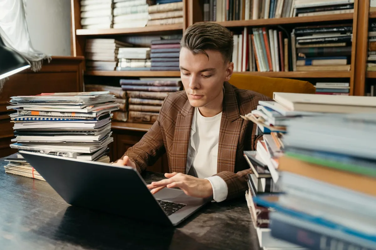 Estudante em biblioteca com laptop e documentos, representando pesquisa acadêmica