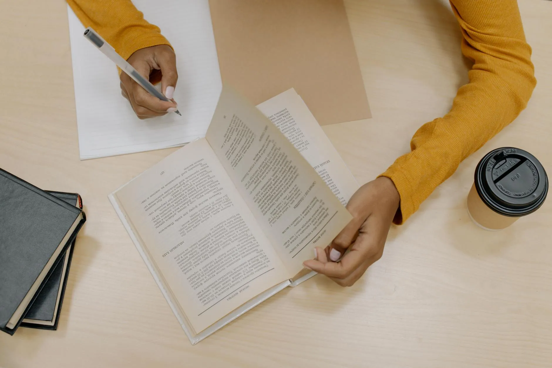 Pilha de livros e cadernos sobre mesa de madeira com caneta e xícara de café ao lado