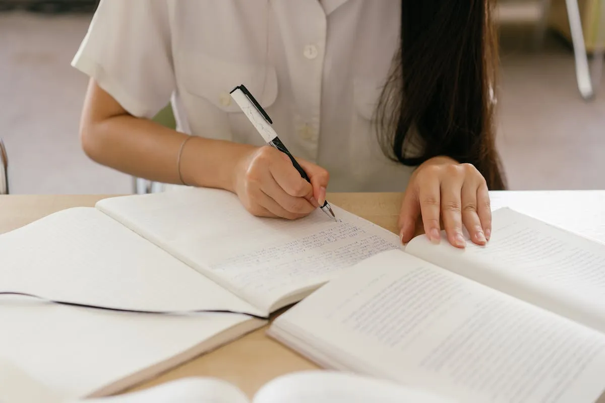Estudante organizando páginas de trabalho acadêmico sobre mesa com computador ao fundo