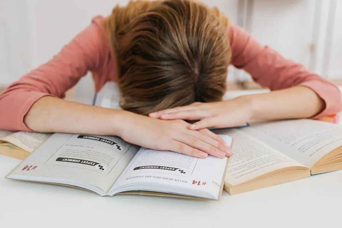 Pessoa com a cabeça apoiada nas mãos em postura de cansaço em frente a livros e cadernos acadêmicos