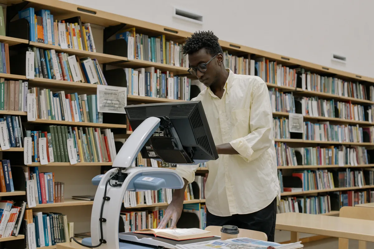 Estudante lendo edital em tela de computador em biblioteca universitária.