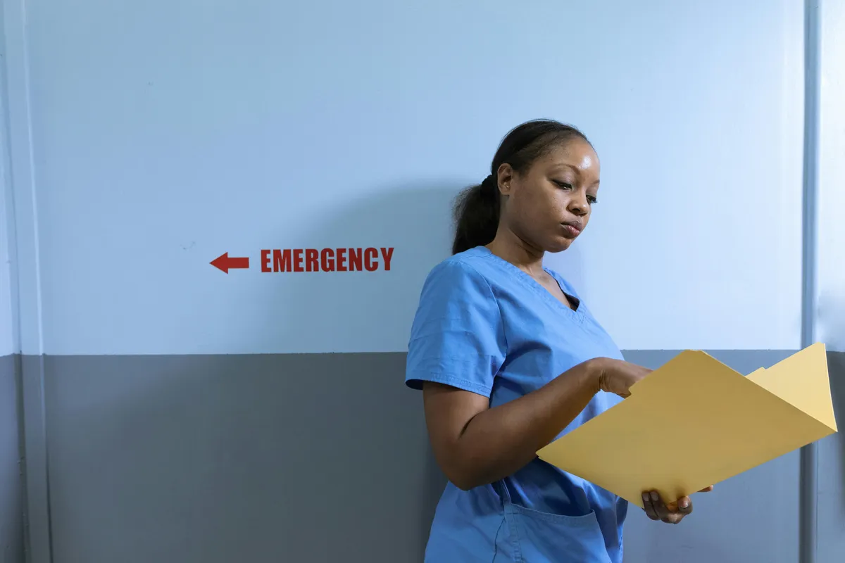 Enfermeira com jaleco branco lendo documentos em mesa de trabalho hospitalar