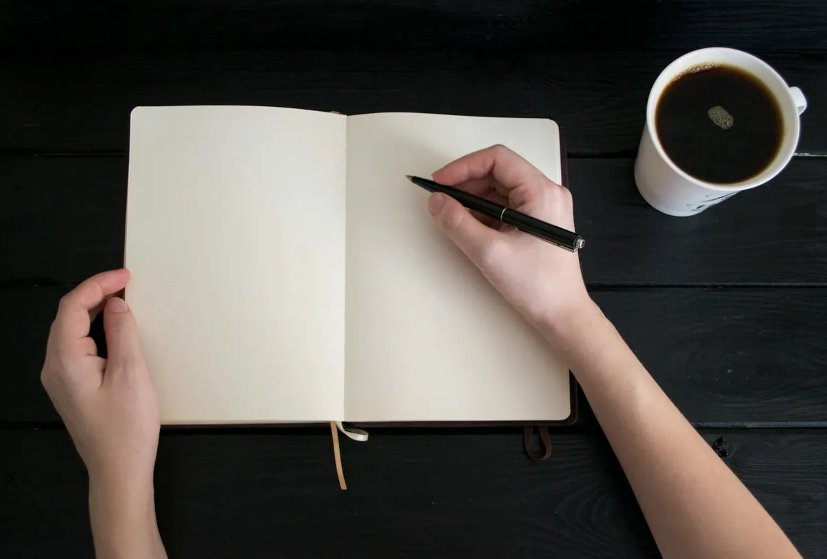 Pesquisadora escrevendo em caderno de diário de pesquisa com café ao lado