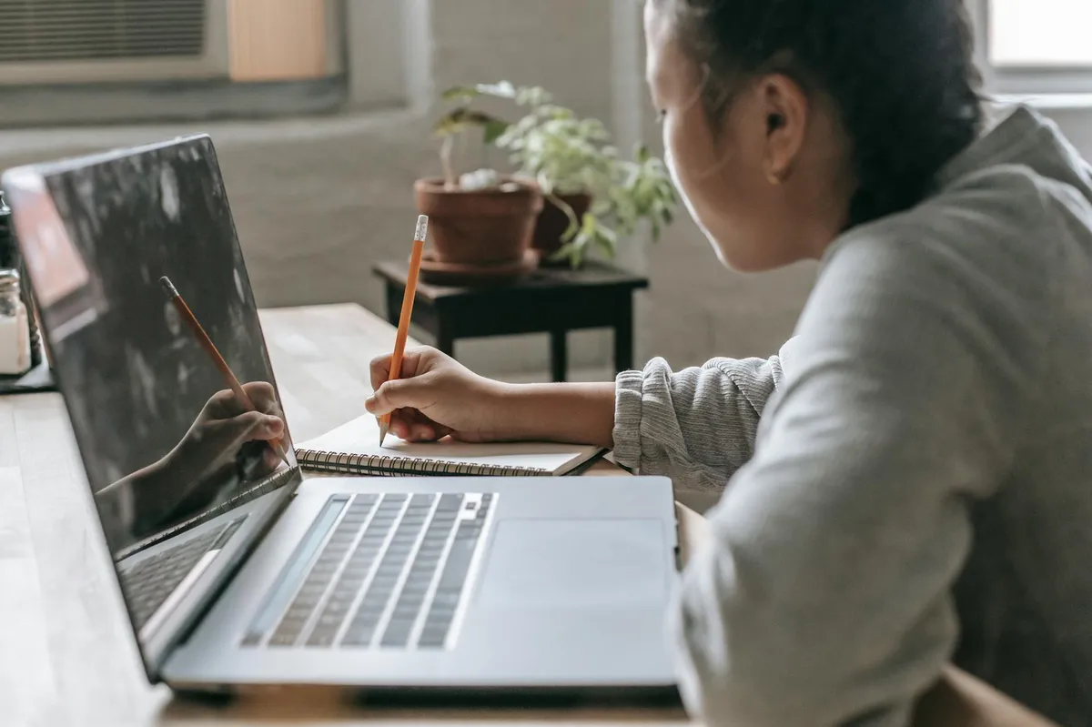 Estudante em frente a laptop com expressão reflexiva, papéis de pesquisa sobre a mesa