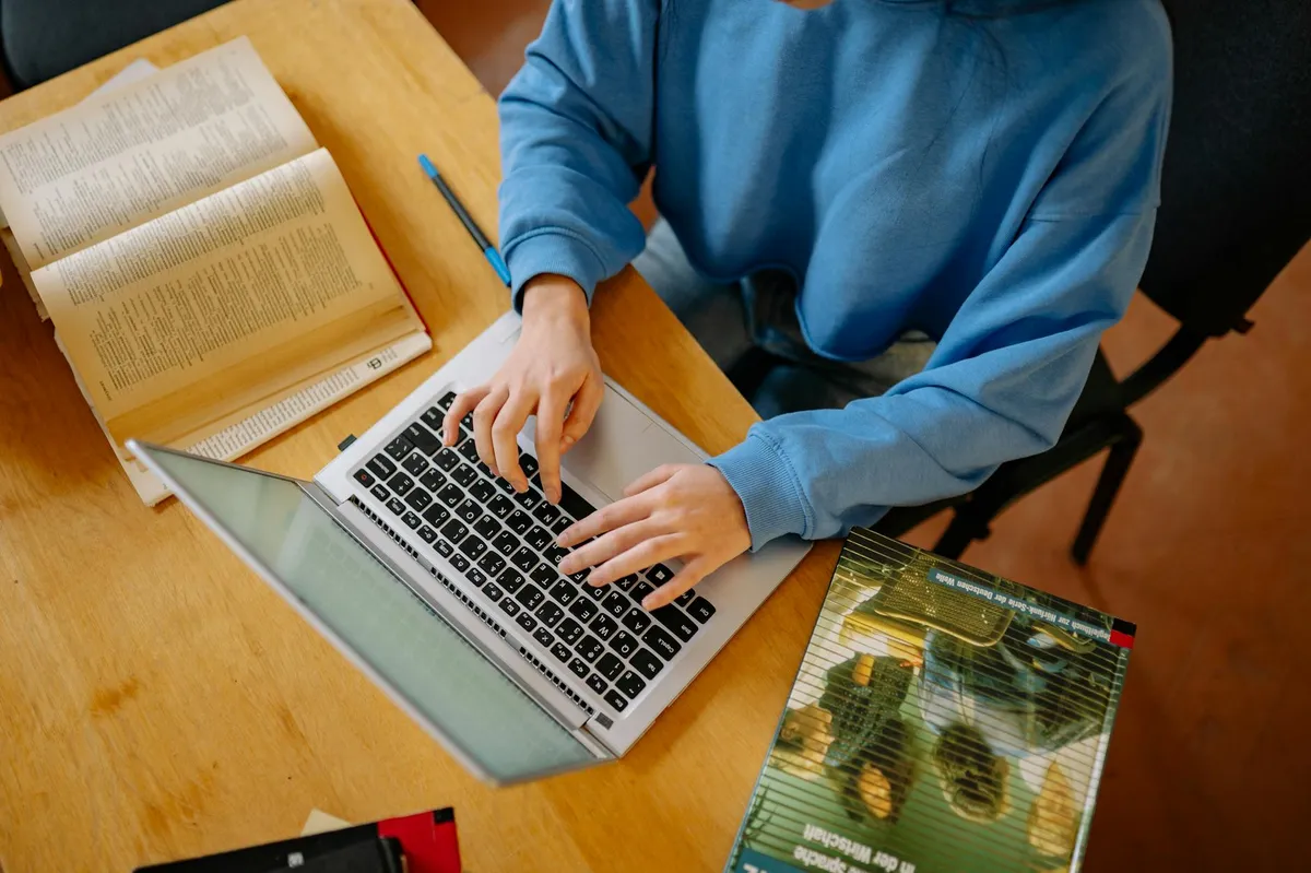 Estudante digitando monografia no laptop com livros ao redor, ambiente de estudo universitário