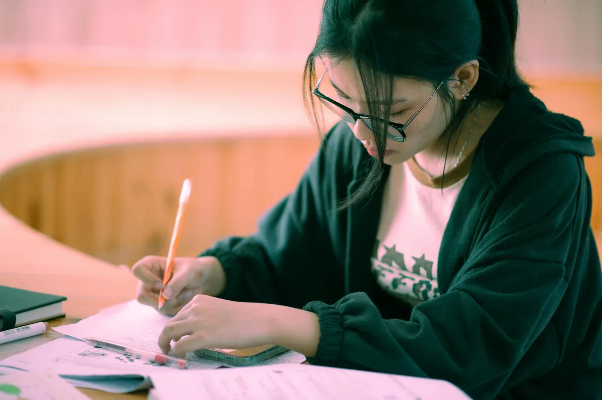Estudante lendo e escrevendo notas ao lado de computador com texto acadêmico na tela