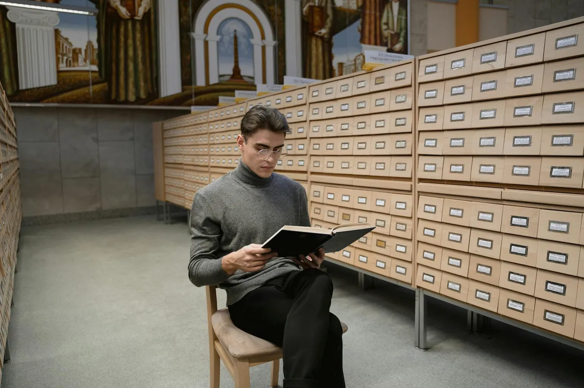 Historiadora pesquisando documentos históricos em biblioteca com computador ao lado