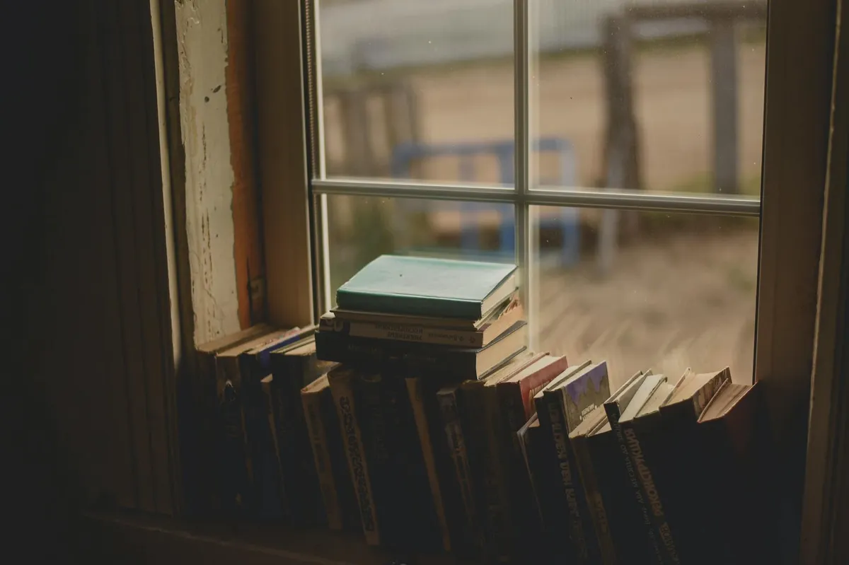 Pessoa olhando pela janela em momento de reflexão, com livros ao fundo