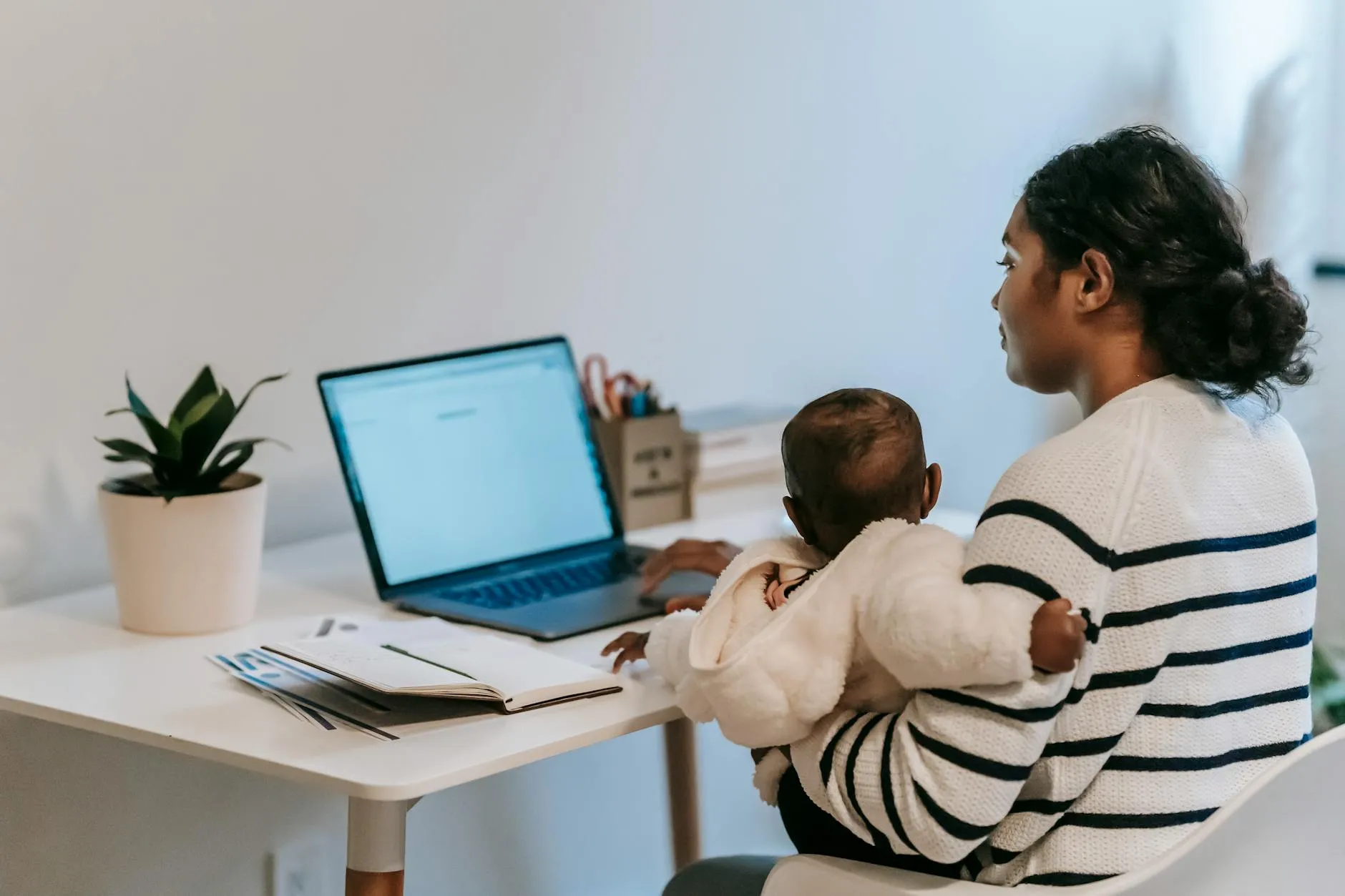 Mulher trabalhando em laptop com bebê no colo em ambiente doméstico iluminado