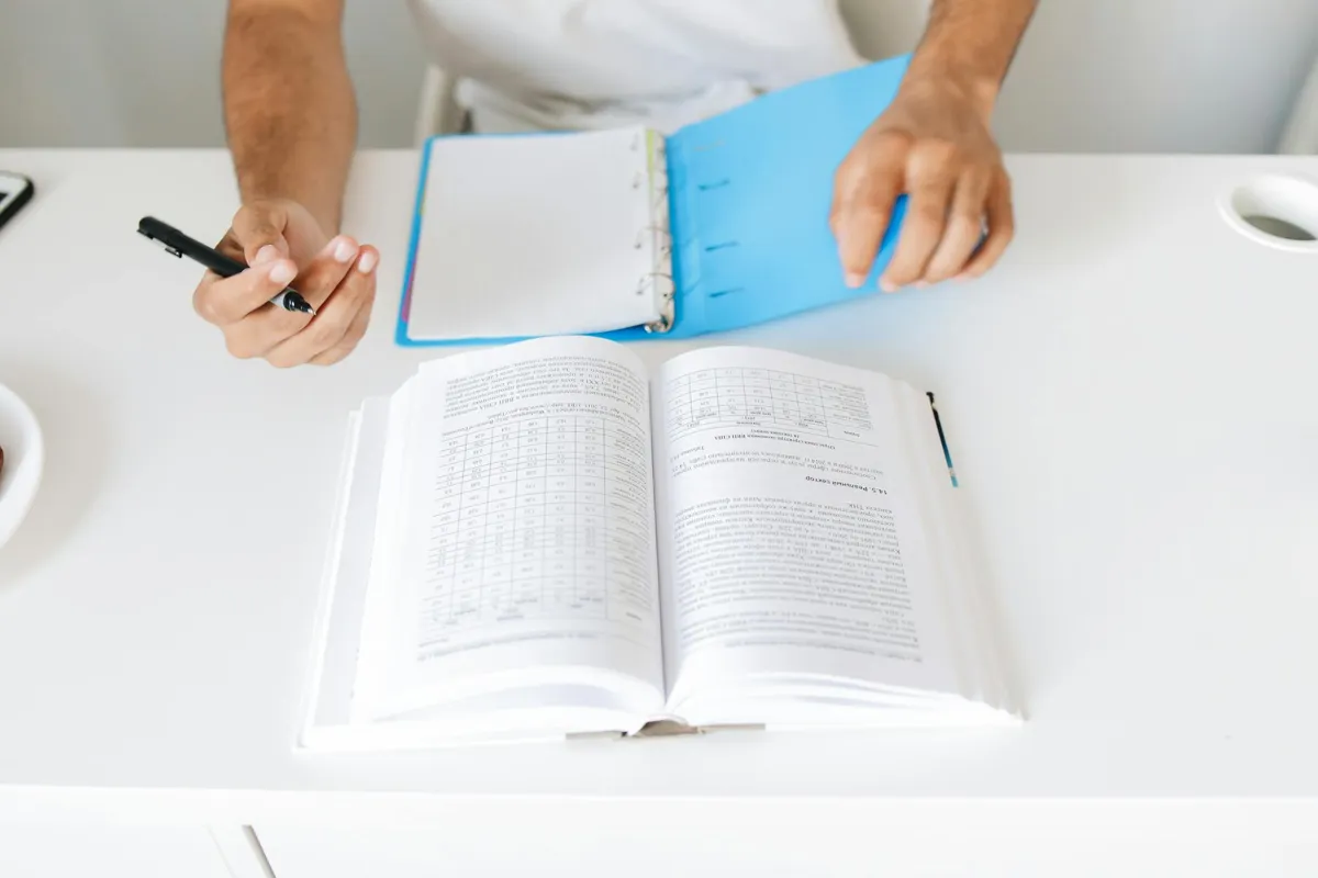 Mesa de estudo com caderno aberto, caneta e livros acadêmicos sobre pesquisa.