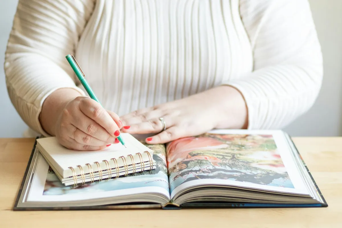 Pessoa escrevendo resenha acadêmica em caderno com livro aberto ao lado