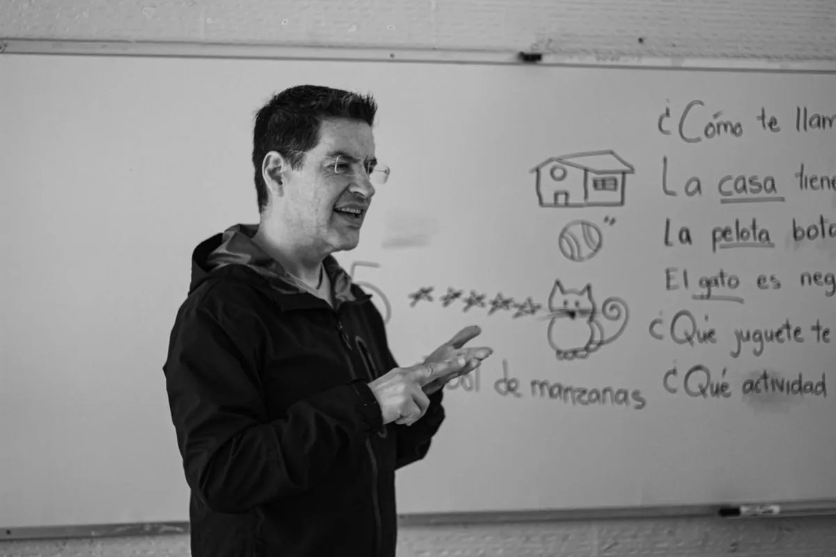 Professor em sala de aula anotando observações em caderno enquanto alunos trabalham em grupo.