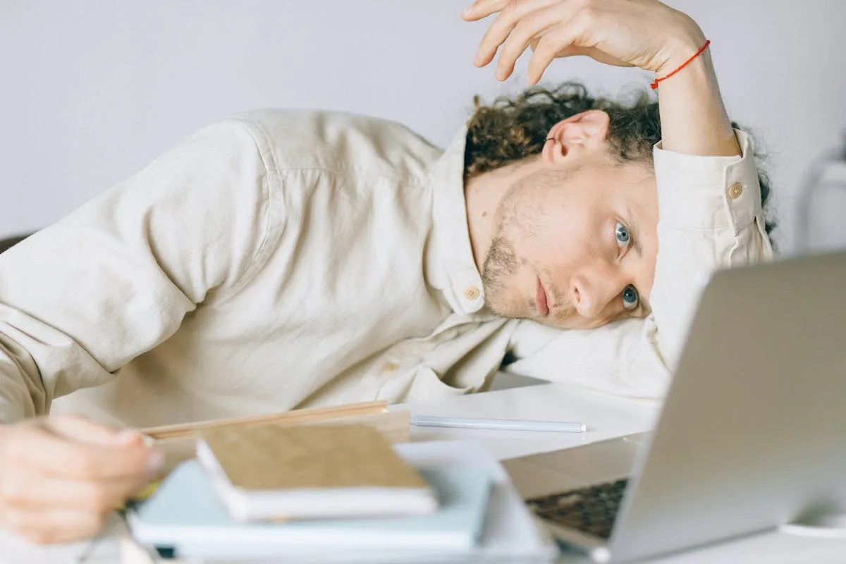 Pessoa cansada sobre mesa com computador, cadernos e papéis de trabalho
