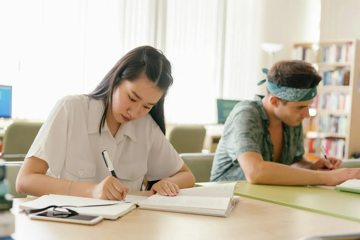 Estudante organizando notas e esquemas em mesa de biblioteca com livros ao fundo.