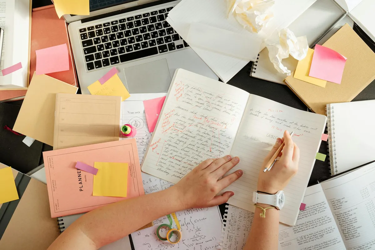 Estudante com caderno aberto e livros ao redor, escrevendo anotações sobre possíveis temas de pesquisa
