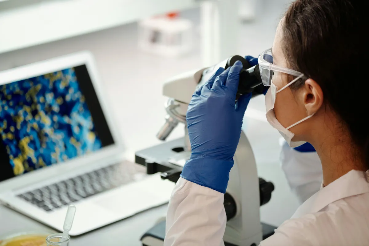 Cientista em jaleco examina amostras com microscópio ao lado de notebook em laboratório de pesquisa
